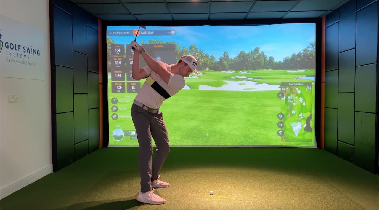 Person playing golf simulation game in a room with a large screen displaying a golf course.